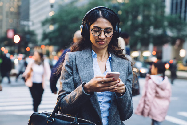 Delighted young brunette ethnic woman in earphones spectacles gray coat and black handbag tapping on smartphone on busy New York street on urban blurred background - 写真・画像