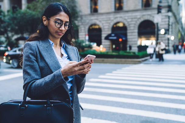 Thoughtful stylish young brunette ethnic woman in round spectacles black handbag and gray coat tapping on smartphone near crosswalk on New York street on blurred background - Foto, imagen