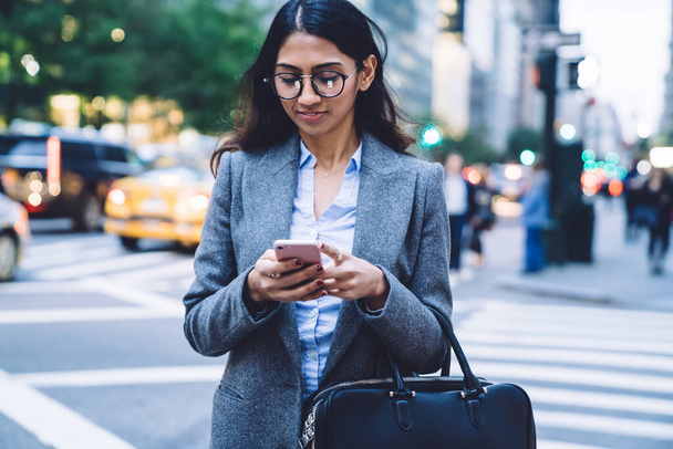 Young beautiful ethnic business woman with black hair and leather bag texting on mobile phone while crossing road in New York City  - Photo, Image