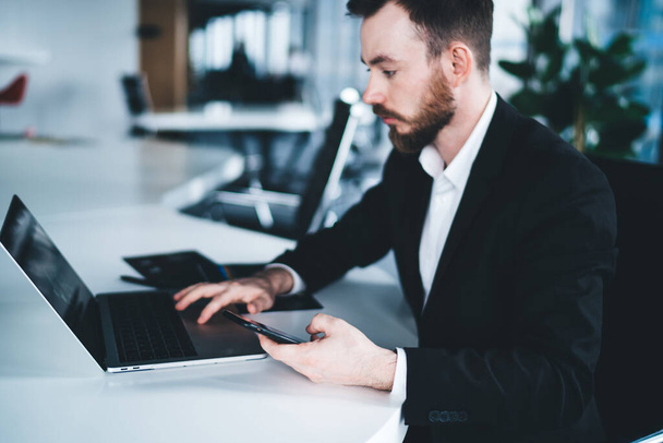 Side view of manager male in formal outfit and beard sitting at desk with documents using laptop while browsing mobile in workspace in company - 写真・画像
