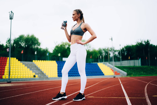 Side view of confident sporty woman in modern sportive wear standing in track holding water bottle working out against stadium seats looking away  - Fotoğraf, Görsel