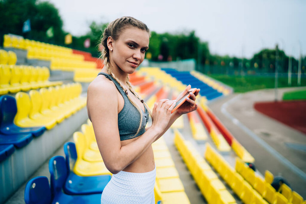 Side view of blond athletic female in sportswear with pigtails messaging on smartphone while standing on empty stadium tribunes and looking at camera on blurred background - Valokuva, kuva
