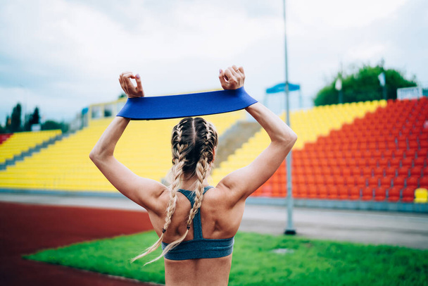 Back view of faceless sporty woman standing and rising up arms while diligently training with blue rubber ribbon at stadium - Foto, Bild