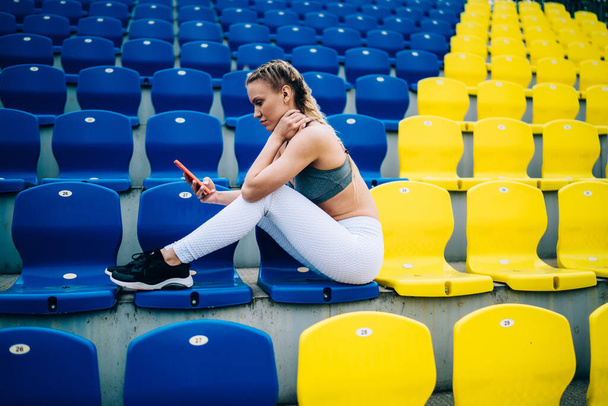 Side view of shapely woman in sneakers and sportswear sitting on stadium seats and resting while surfing internet using orange smartphone - Photo, image
