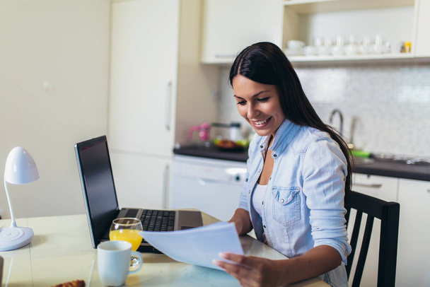 Attractive young woman working with laptop computer and documents while sitting at the kitchen. - Φωτογραφία, εικόνα