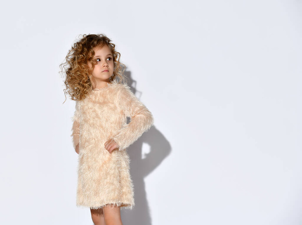 Little blonde curly kid in fluffy dress and shoes. She has put her hands on hips, posing standing sideways isolated on white - 写真・画像