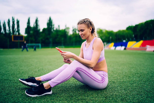 Side view of adult sporty female in modern sportive wear browsing cellphone while sitting on green grass of stadium field - Фото, изображение