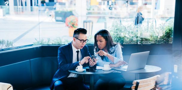 Young multiracial woman and man sharing mobile phone while working in team on new project sitting at table in cafe - Photo, Image