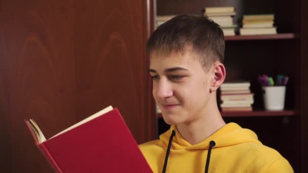Mladý student doma čte knihu na pozadí knihovny s učebnicemi. Ve žluté mikině. Detailní záběr. Online distanční vzdělávání - Záběry, video