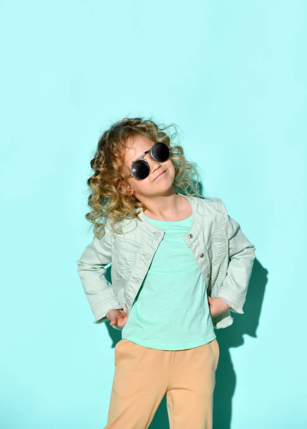 Cute little curly girl with black non-transparent glasses on face stands raising one hand pointing two fingers up. Studio shot isolated on turquoise - Foto, imagen