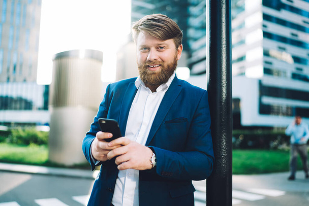 Half length portrait of cheerful successful man in formal wear holding cellphone gadegt in hands and smiling at camera during time in financial district, positive bearded boss with smartphone - Photo, image