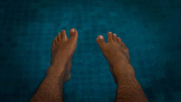 Un hombre relajado con los pies en la piscina del hotel durante el verano. Concepto oscuro y malhumorado. Vista selectiva
. - Foto, imagen