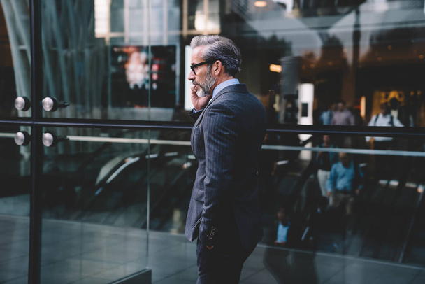 Side view of businessman with beard in fitting well dark business suit talking on phone near glass wall of office building putting hand in pocket in New York - Foto, Imagen