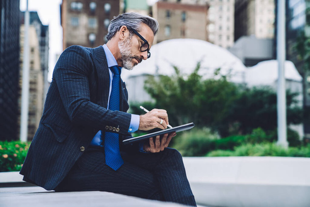 Side view of middle aged businessman in glasses with legs crossed working on tablet with stylus while sitting on bench against background of New York buildings - Foto, Imagem