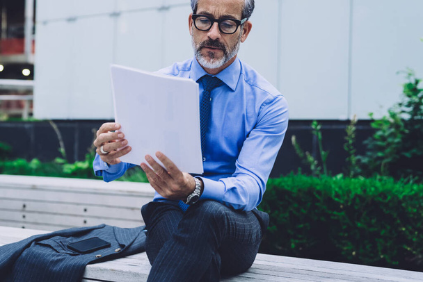 Handsome concentrated middle aged man in blue shirt and glasses analyzing papers and sitting on wooden bench with blazer on New York street - Foto, Imagem