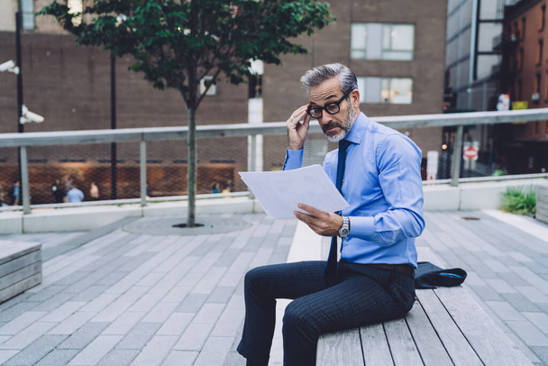 Side view of middle aged man in business suit correcting glasses while sitting on wooden bench and analyzing papers on New York street - Foto, afbeelding
