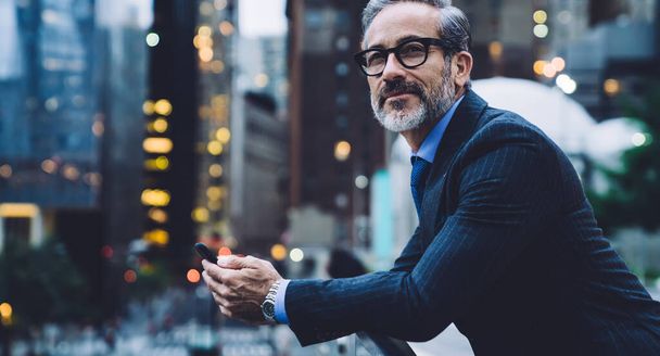 Middle aged man in glasses and business suit holding phone and leaning on glass fence against background of New York street at evening - Photo, Image