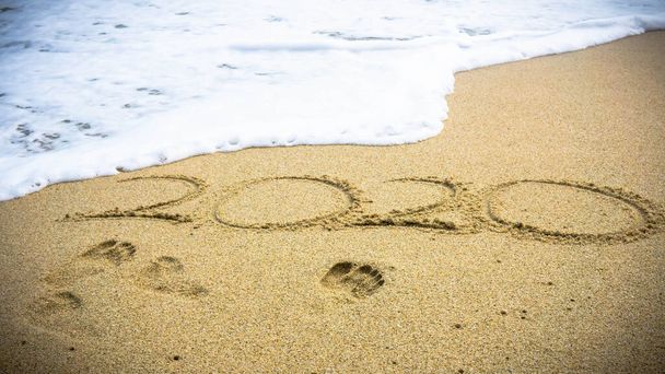 Feliz Año Nuevo 2020, letras en la playa con olas marinas. Números 2020 año en la orilla del mar, mensaje escrito a mano sobre la arena en el hermoso fondo de la playa. La imagen está un poco fuera de foco. Huella de arena
. - Foto, imagen