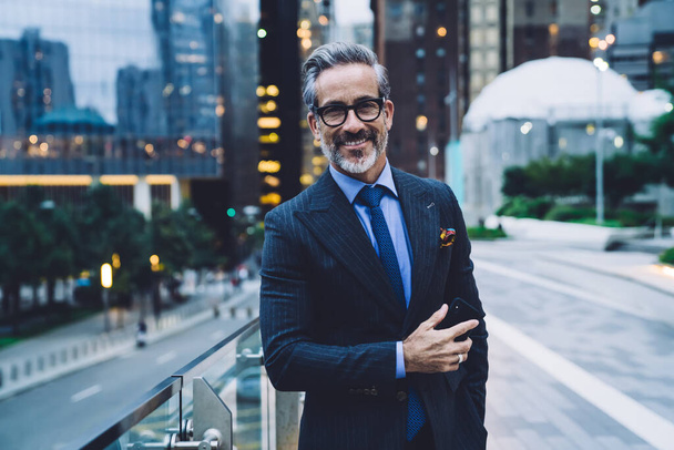 Handsome bearded middle aged man in glasses and business suit holding phone and standing against evening New York street while looking at camera - Foto, immagini