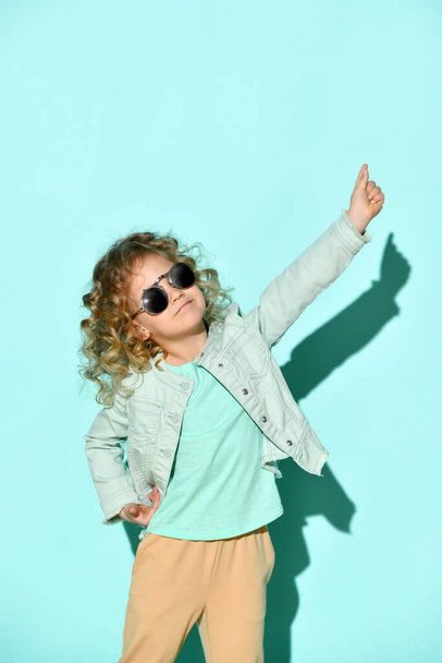 Cute little curly girl with black non-transparent glasses on face stands raising one hand pointing two fingers up. Studio shot isolated on turquoise - Zdjęcie, obraz