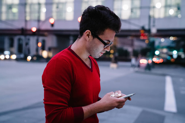 Side view of stylish young man in glasses and red sweater browsing smartphone while standing on city street in twilight - Foto, Bild