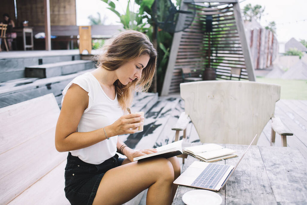 Beautiful focused woman in casual clothing reading book and drinking coffee while sitting in cafe on wooden table with laptop and notebook - Foto, Imagen