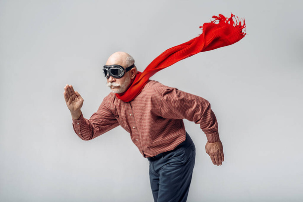 Elderly man in a red scarf and pilot glasses flying like a superman, grey background. Cheerful mature senior looking at camera in studio - Photo, Image
