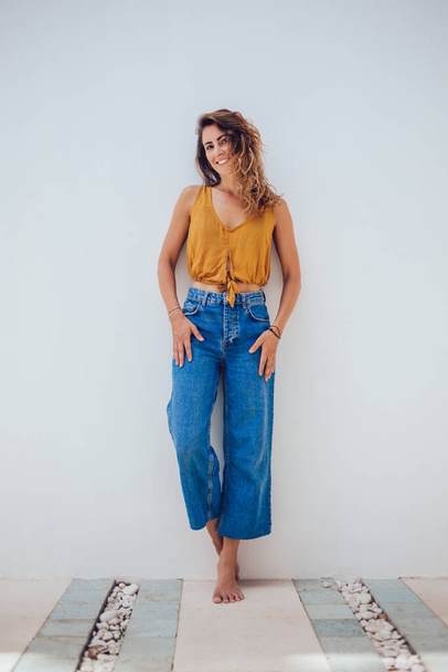 Peaceful subtle bronzed shoeless woman in yellow summer top and jeans posturing by wall on white background and looking at camera - Foto, imagen