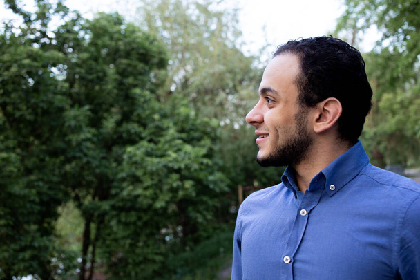 Úzký profil mladého muže ze Středního východu. Detailní portrét usměvavého muže z Egypta v modré košili v parku. Stromy na pozadí. - Fotografie, Obrázek