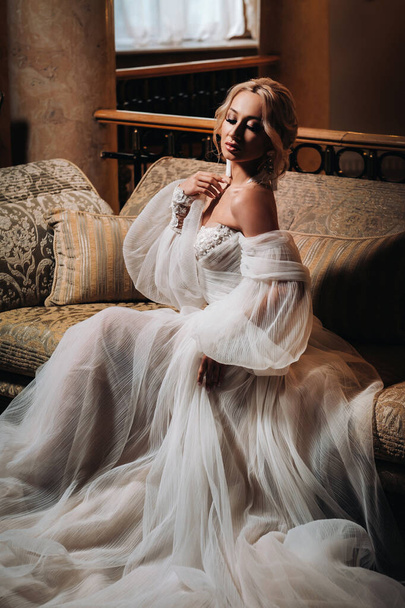 A beautiful bride is sitting on a luxurious sofa. Beautiful model girl in a white wedding dress. Female portrait of a nice lady. Woman with hair. - Zdjęcie, obraz