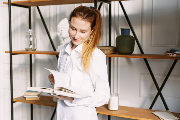 Menina bonita estudar em casa. Uma jovem está lendo um livro na frente de uma estante elegante e moderna
. - Foto, Imagem