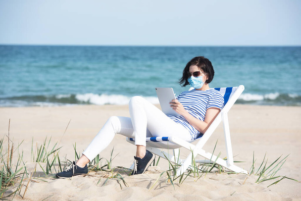Junge Frau in Maske sitzt mit Laptop auf leerer Küste, Schutzausrüstung während Coronavirus covid19. Soziales Distanzkonzept. - Foto, Bild
