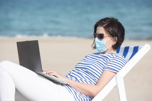 Maskeli genç bir kadın boş sahilde dizüstü bilgisayarla oturuyor. Koronavirüs sırasında koruyucu ekipmanlar. Sosyal uzaklık kavramı. - Fotoğraf, Görsel