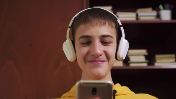 Young man student in white wireless headphones plays and writes in a smartphone on the background of a bookcase with textbooks and school supplies. Close-up. Online learning to coronavirus quarantine - Felvétel, videó