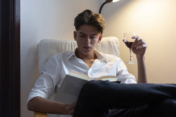 Portrait of young man in white shirt sitting drinking wine and reading a book at home. - Photo, Image