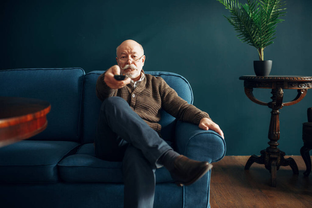 Elderly man watching TV on couch in home office. Bearded mature senior poses in living room, old age businessman - Foto, imagen