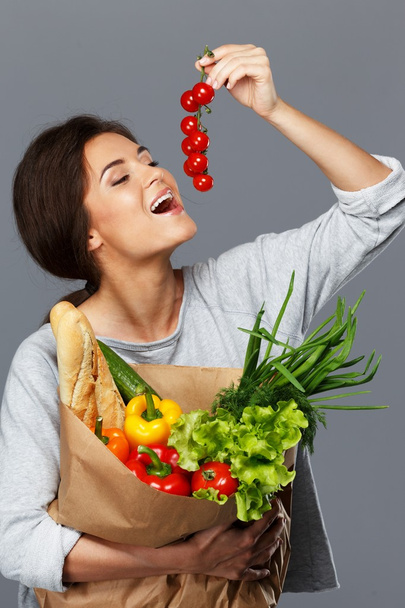 lachende brunette vrouw met kruidenier zak vol met verse groenten en cherry tomaten - Foto, afbeelding