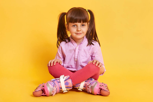 Closeup portrait of calm little girl sitting on floor with crossed legs, wearing casual attire and rolling skates, having dark hair and two ponytails, looks at camera, keeping palms on knees, has rest - Photo, image