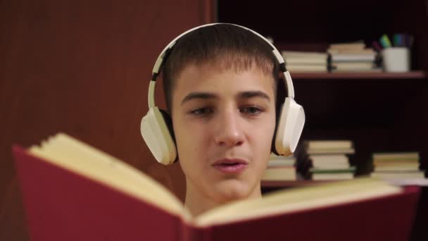 Giovane studente legge ad alta voce un libro in cuffia sullo sfondo di una libreria di legno primo piano. Coronavirus quarantena apprendimento online
 - Filmati, video