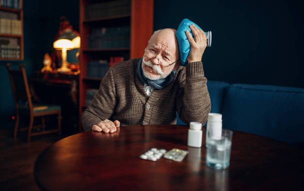 Ο άρρωστος ηλικιωμένος βάζει πάγο στο κεφάλι του στο γραφείο του σπιτιού, ασθένειες που σχετίζονται με την ηλικία, πονοκέφαλος. Ο μεγαλύτερος είναι άρρωστος και νοσηλεύεται στο σπίτι του. - Φωτογραφία, εικόνα