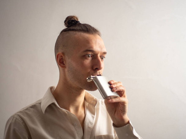 Νεαρός Ξανθός Καυκάσιος άντρας με λευκό πουκάμισο πίνει λίγο αλκοόλ στο ασημένιο του Hip Flask στην καραντίνα του Coronavirus. - Φωτογραφία, εικόνα