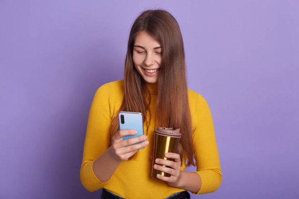 Closeup portrait of smiling woman with coffee or tea in hands, holding smart phone and checking social networks, enjoying hot beverage, looking at device's display and smiles. Modern technology. - Foto, Imagen