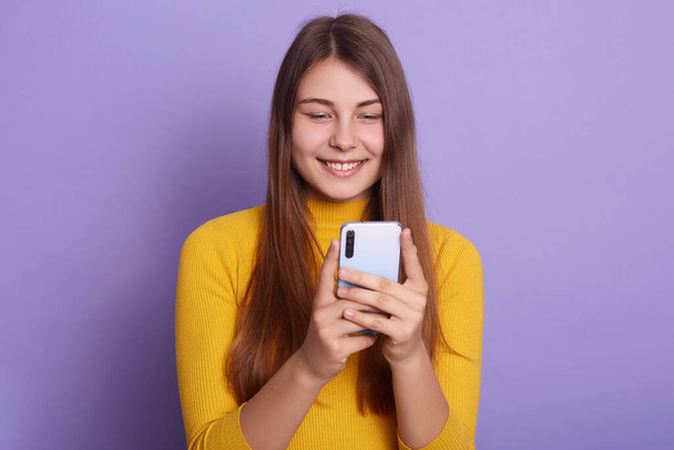 Pretty attractive woman with positive facial expression, wearing casual attire, posing isolated over lilac studio background, holding mobile phone in hands, looking at screen, checking social network. - 写真・画像
