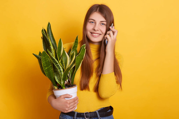 Studio shot of happy pretty female dresses casual attire, talking to somebody via phone, holding in hands white flower pot with potted sansevieria, stands against yellow wall, expresses positive. - Φωτογραφία, εικόνα