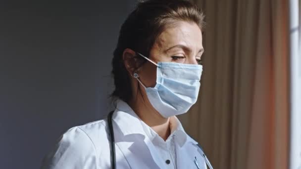 Charismatic woman doctor with a tired eyes and wearing a protective mask looking through the hospital window closeup to the camera - Video