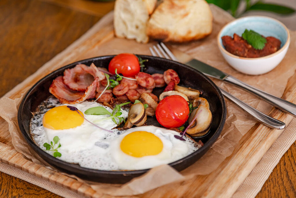 English breakfast. Fried eggs with fried sausages, bacon, tomatoes and mushrooms. On a wooden with bean paste. - Photo, Image