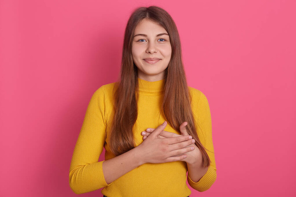 Studio shot of young Caucasian lady posing indoor against pink background, keeping hands on chest, has grateful facial expression, wearing casual attire, looking at camera, being thankful to someone. - 写真・画像