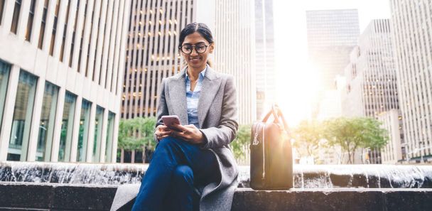 Χαμογελώντας εθνοτική επιχειρηματίας κυρία με κομψό παλτό και γυαλιά ηλίου κάθεται με τσάντα στην άκρη του σιντριβανιού και χρησιμοποιώντας το κινητό τηλέφωνο στο δρόμο της Νέας Υόρκης - Φωτογραφία, εικόνα