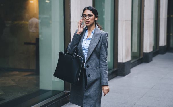 Attractive business lady in elegant outfit and eyeglasses holding leather bag having phone call while talking at street of New York city - Fotoğraf, Görsel