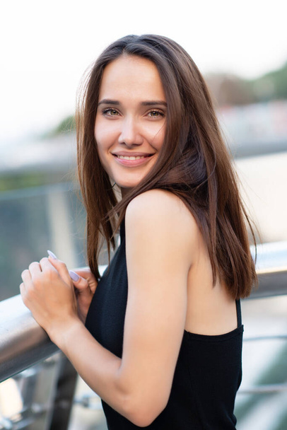 Young pretty likable cheerful woman posing summer city outdoor in Kyiv, Ukraine. Beautiful self-confident girl with long brown hair posing on Dnipro river bank enjoing her life, urban lifestyle - Foto, afbeelding